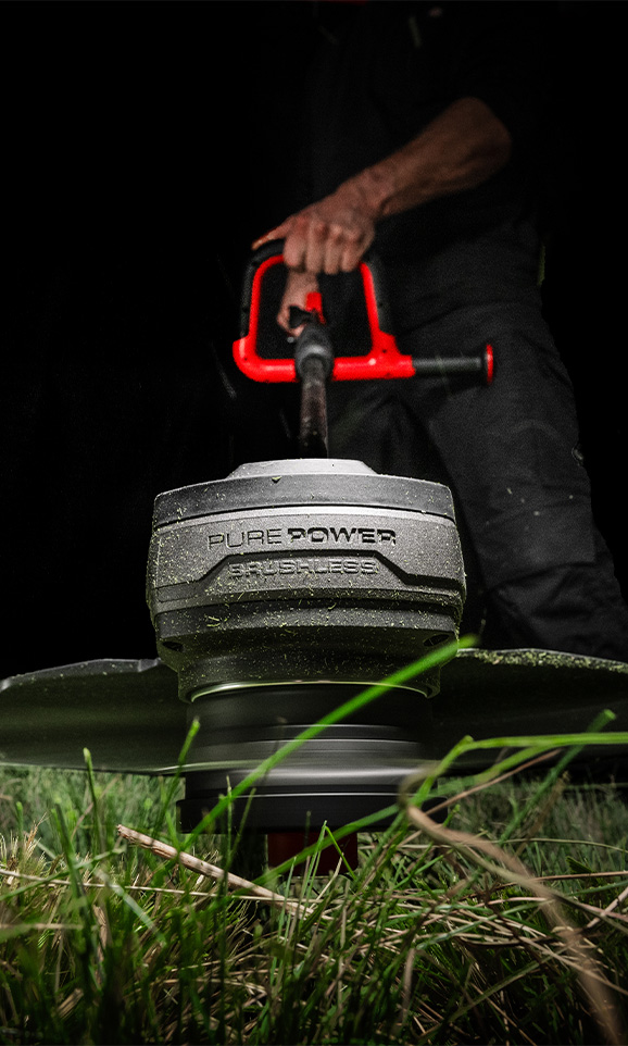 A red Einhell cordless grass trimmer is operated by a person in the background, with the focus on the spinning cutting head.