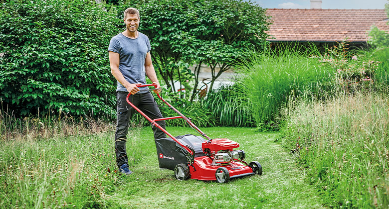 A man standing on a strip of mown grass in a meadow, a petrol lawnmower in front of him.